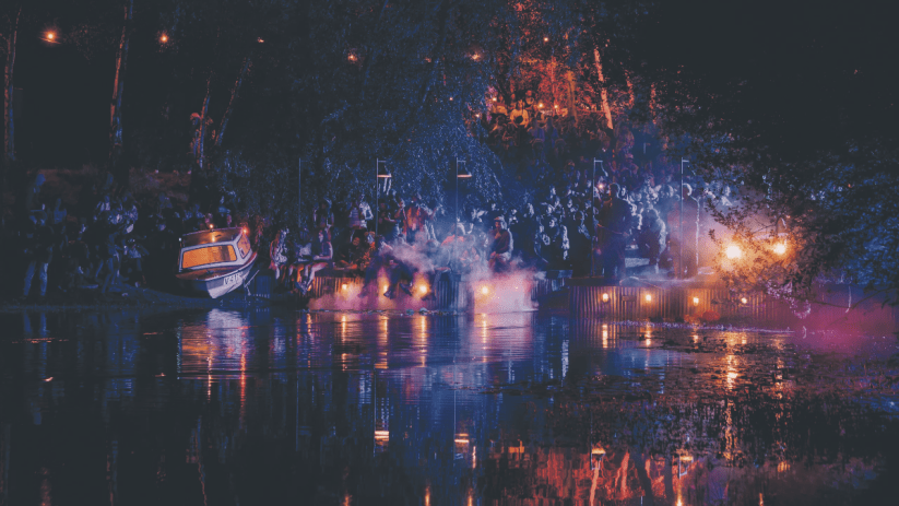 Lost Village festival at night: crowd gathered by a lake, lit by lanterns and colorful lights, reflecting on the water.