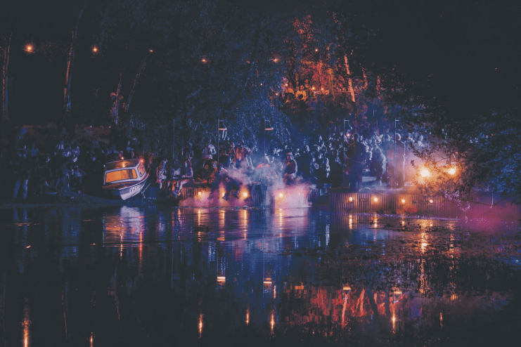 Lost Village festival at night: crowd gathered by a lake, lit by lanterns and colorful lights, reflecting on the water.