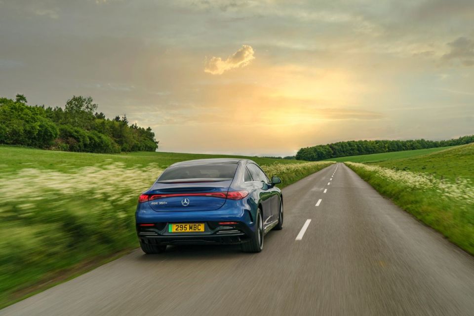 Blue Mercedes-Benz EQE 350+ electric sedan driving on a country road at sunset, surrounded by green fields.
