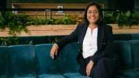 Neetu Mistry, a smiling woman with dark hair, wearing a navy suit and white shirt, sitting on a teal couch