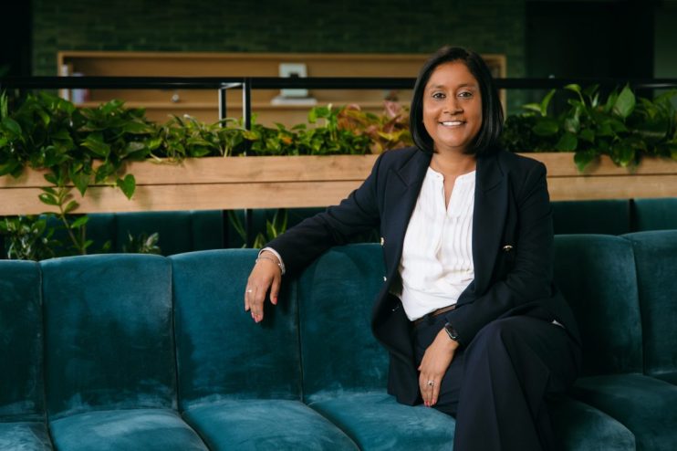 Neetu Mistry, a smiling woman with dark hair, wearing a navy suit and white shirt, sitting on a teal couch