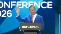 Nigel Farage at a Reform UK conference, gesturing from a podium with the party logo and CONFERENCE 2026 in background