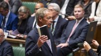 Nigel Farage in Parliament, addressing a female clerk holding a document, with other parliamentarians in the background.