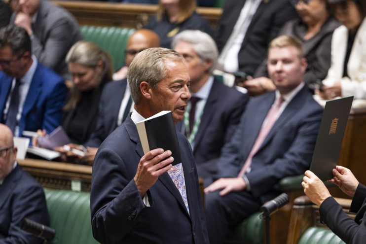 Nigel Farage in Parliament, addressing a female clerk holding a document, with other parliamentarians in the background.