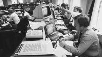Office worker in the 1990s, surrounded by vintage computer monitors, calculators, and paper documents.