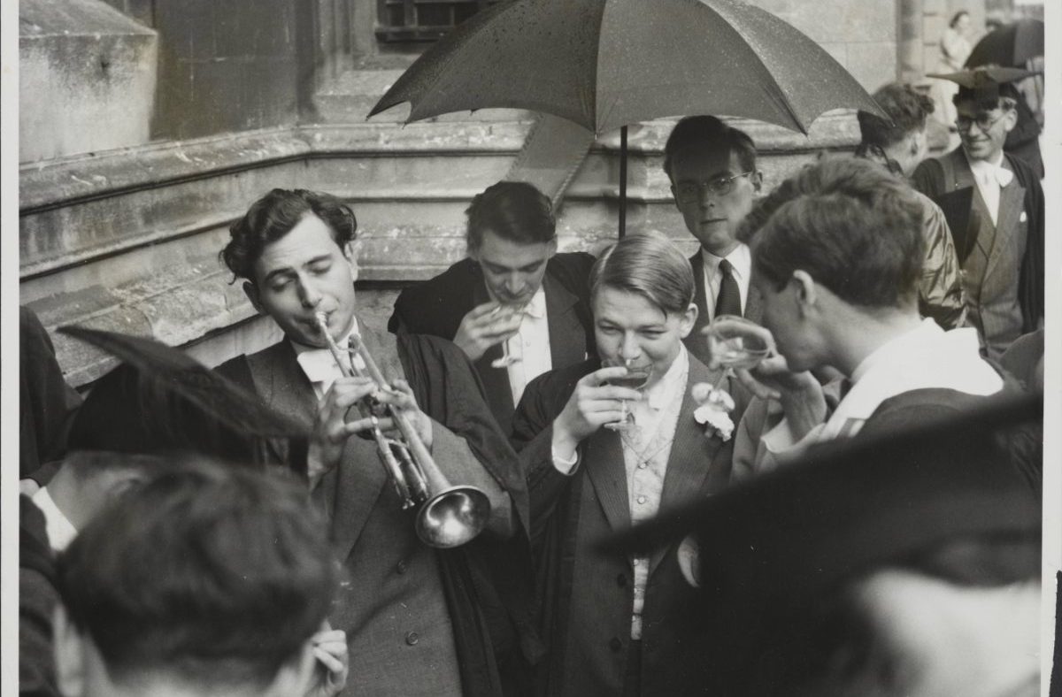 Oxford University students in academic gowns, one playing a trumpet, others drinking under an umbrella.
