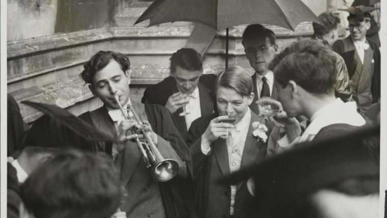 Oxford University students in academic gowns, one playing a trumpet, others drinking under an umbrella.