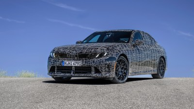 Camouflaged BMW sedan prototype with black and white pattern on an asphalt road under a clear blue sky