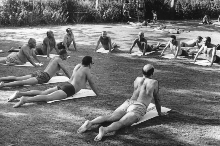 Men in swim trunks practicing yoga in a park, performing cobra pose on mats.