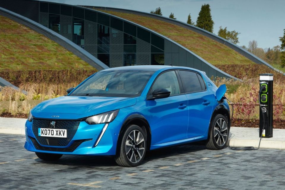 Blue Peugeot 208 electric car charging at an outdoor station with a modern building and green roof in the background.