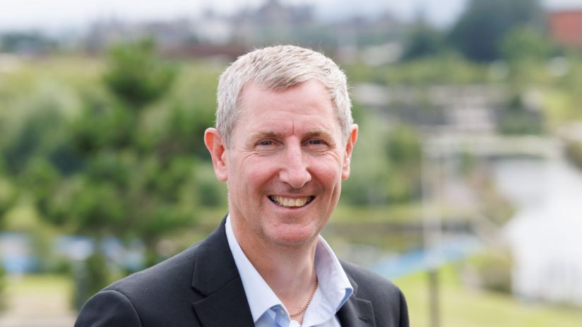 Smiling man in a suit jacket and light blue shirt, with a blurred cityscape and greenery in the background
