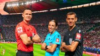 Three football referees, two men and one woman, stand before a large screen displaying a stadium and UEFA Strauss logos.