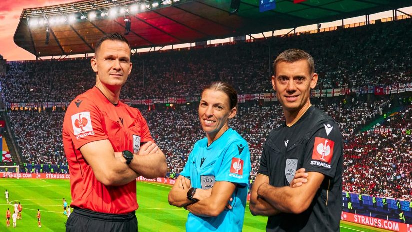 Three football referees, two men and one woman, stand before a large screen displaying a stadium and UEFA Strauss logos.