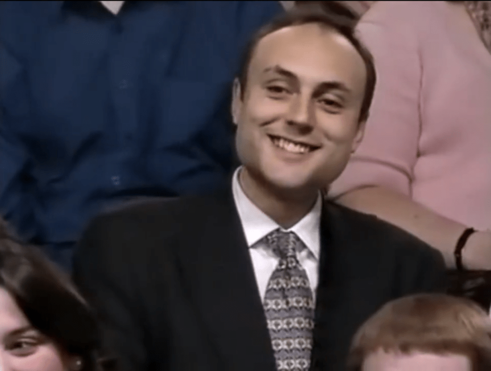 Smiling man in a suit and patterned tie, seated in a group setting, possibly an audience or panel