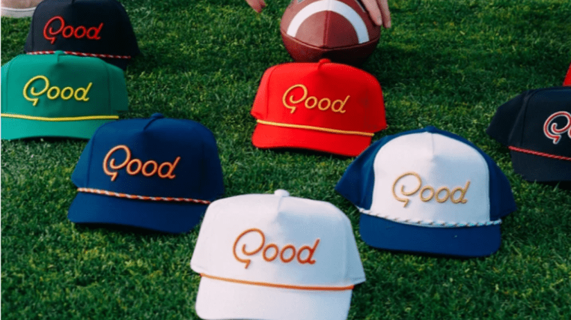 Assortment of colorful Good embroidered baseball caps on green grass with a football and persons hand.