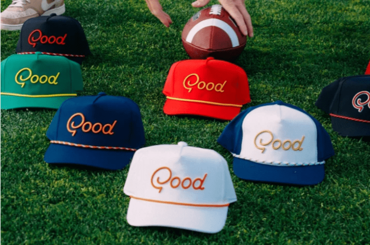 Assortment of colorful Good embroidered baseball caps on green grass with a football and persons hand.