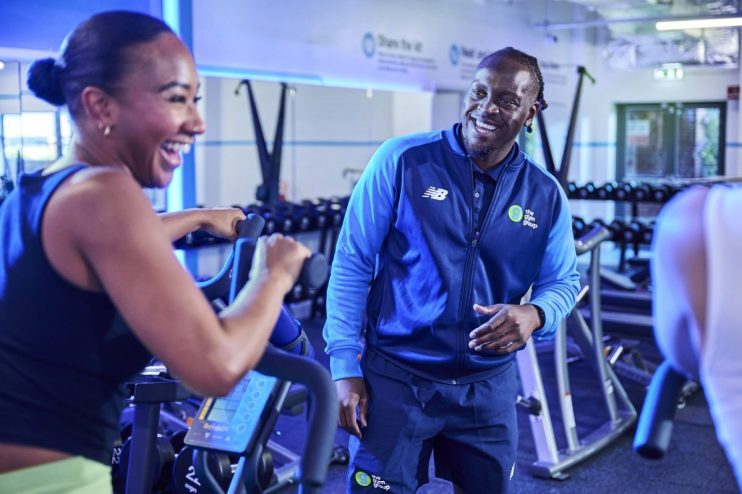 Man in The Gym Group uniform smiling at a woman on an exercise bike.