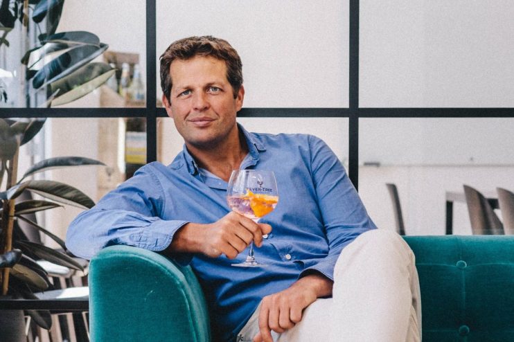 Tim Warrillow seated on a teal sofa, holding a drink with orange slices, in a modern office.