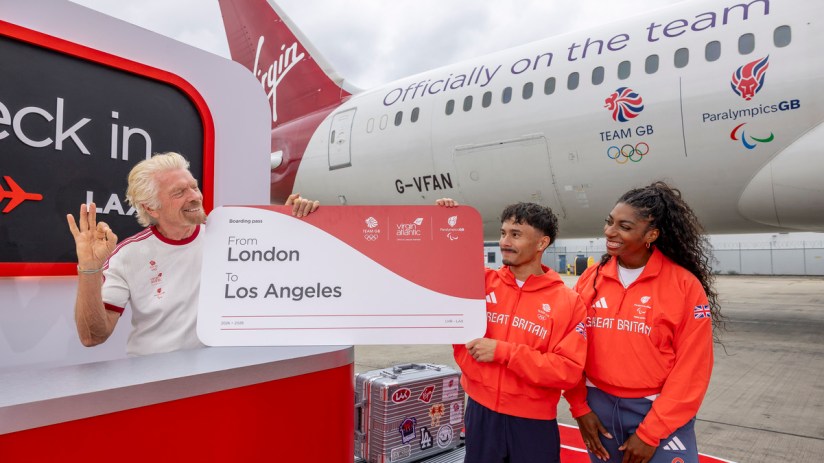 Richard Branson with Team GB athletes holding a London to Los Angeles boarding pass by a Virgin Atlantic plane.