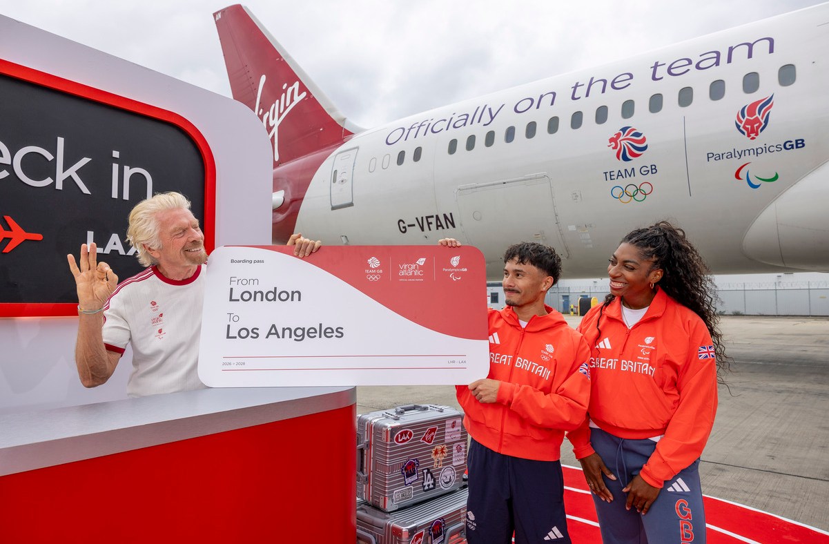 Richard Branson with Team GB athletes holding a London to Los Angeles boarding pass by a Virgin Atlantic plane.