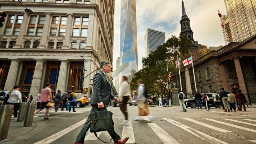 Businessman crossing a Wall Street intersection with One World Trade Center and Trinity Church in the background.