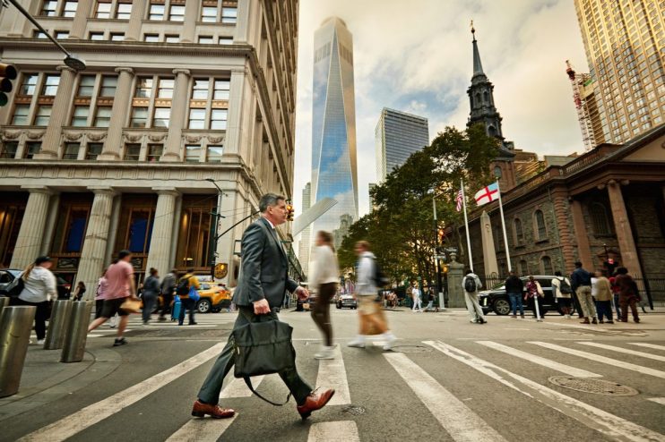 Businessman crossing a Wall Street intersection with One World Trade Center and Trinity Church in the background.