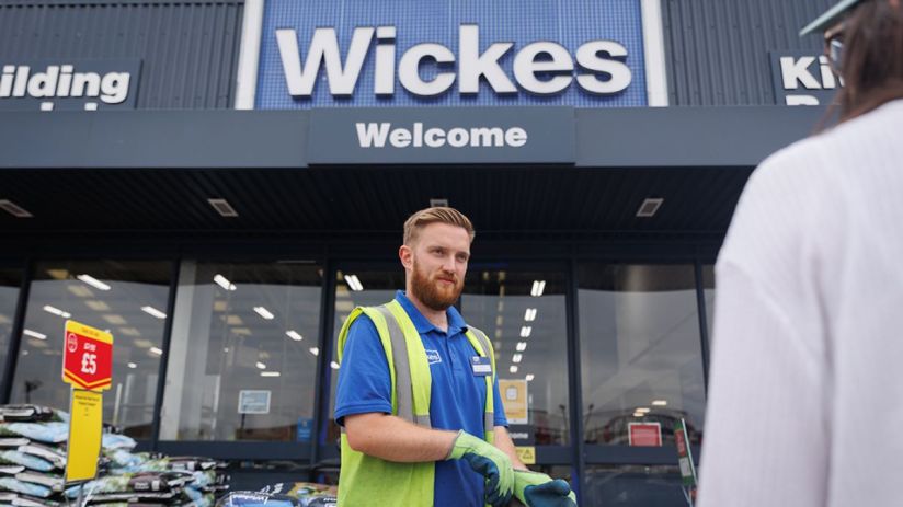 Wickes employee in a high-vis vest and gloves standing outside a store entrance with bags of soil for sale.