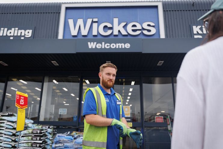 Wickes employee in a high-vis vest and gloves standing outside a store entrance with bags of soil for sale.