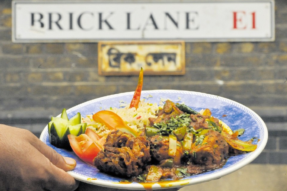 Curry dish with rice, vegetables, and a red chili on a plate held by a hand, Brick Lane sign in background