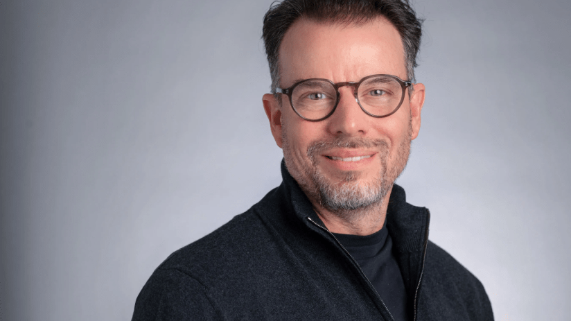 Chris Rokos, a man with glasses and a beard, smiling in a dark zipped sweater against a grey background.