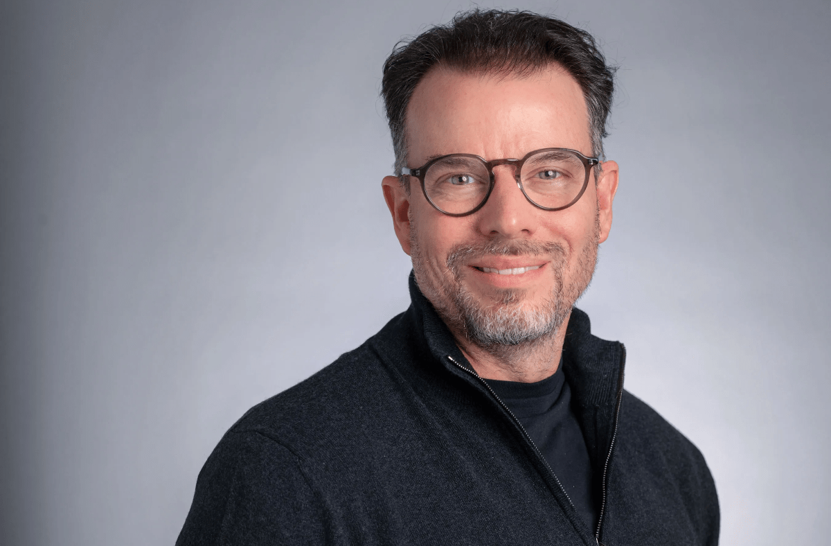Chris Rokos, a man with glasses and a beard, smiling in a dark zipped sweater against a grey background.