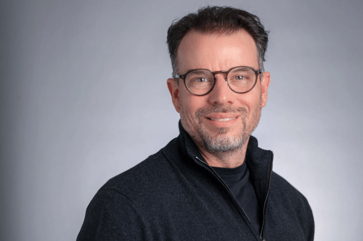 Chris Rokos, a man with glasses and a beard, smiling in a dark zipped sweater against a grey background.