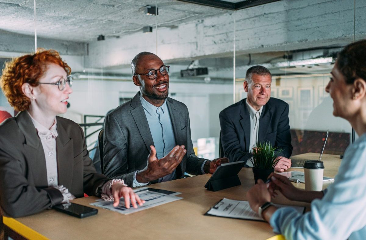 Diverse business professionals in a modern office meeting, discussing strategies with a tablet and documents.