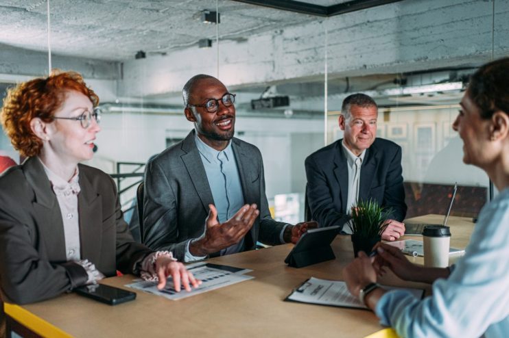 Diverse business professionals in a modern office meeting, discussing strategies with a tablet and documents.