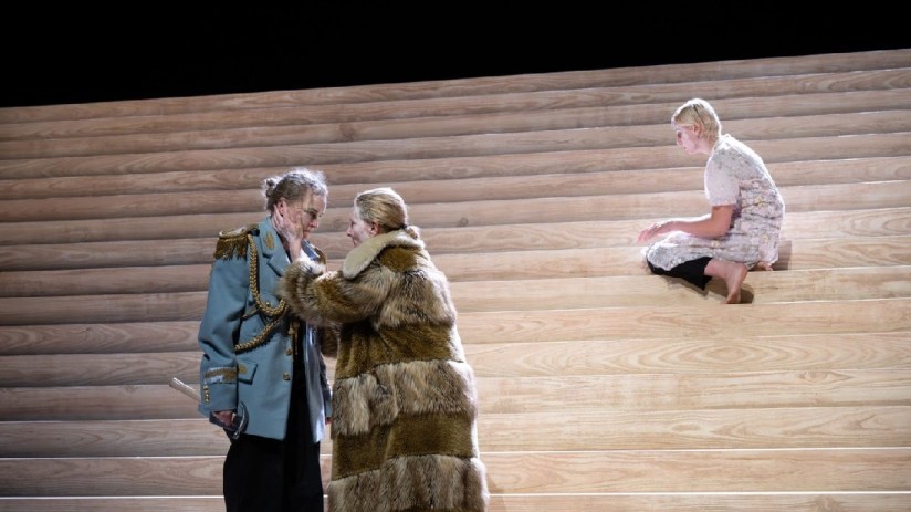 Three actors on a minimalist wooden stage, two embracing, one sitting apart.