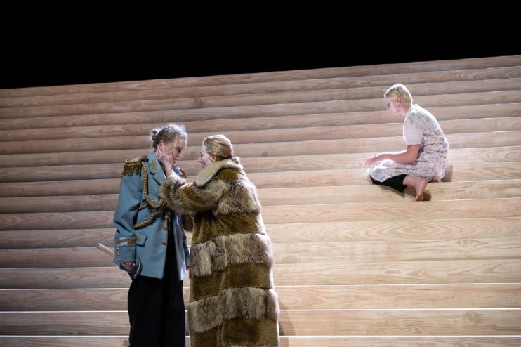 Three actors on a minimalist wooden stage, two embracing, one sitting apart.