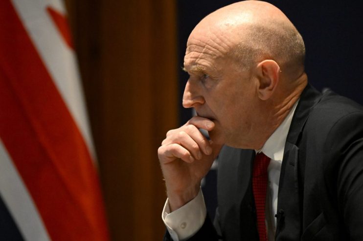 Patrick Healey in a suit with a red tie, looking contemplatively to the left, with an American flag behind him.