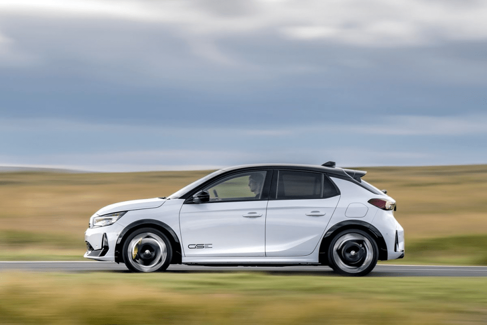 White 2023 Opel Corsa-e GSE electric car driving on a rural road with motion blur.