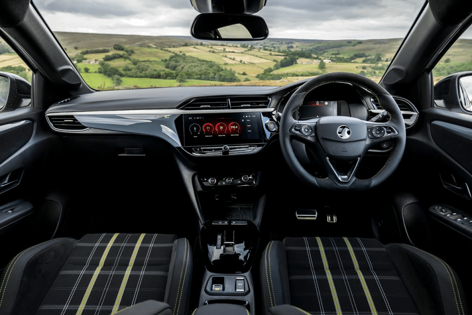 Interior view of a modern car dashboard with a digital display, steering wheel, and plaid seats, overlooking a scenic land...