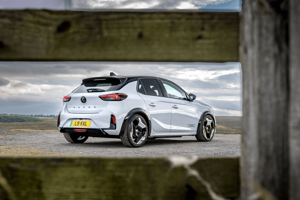 White Vauxhall Corsa hatchback with black roof, seen from the rear, on a gravel road with a cloudy sky