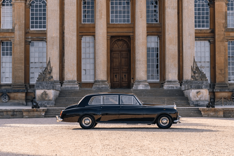 Black vintage luxury car parked on gravel in front of a grand stone building with columns and large windows