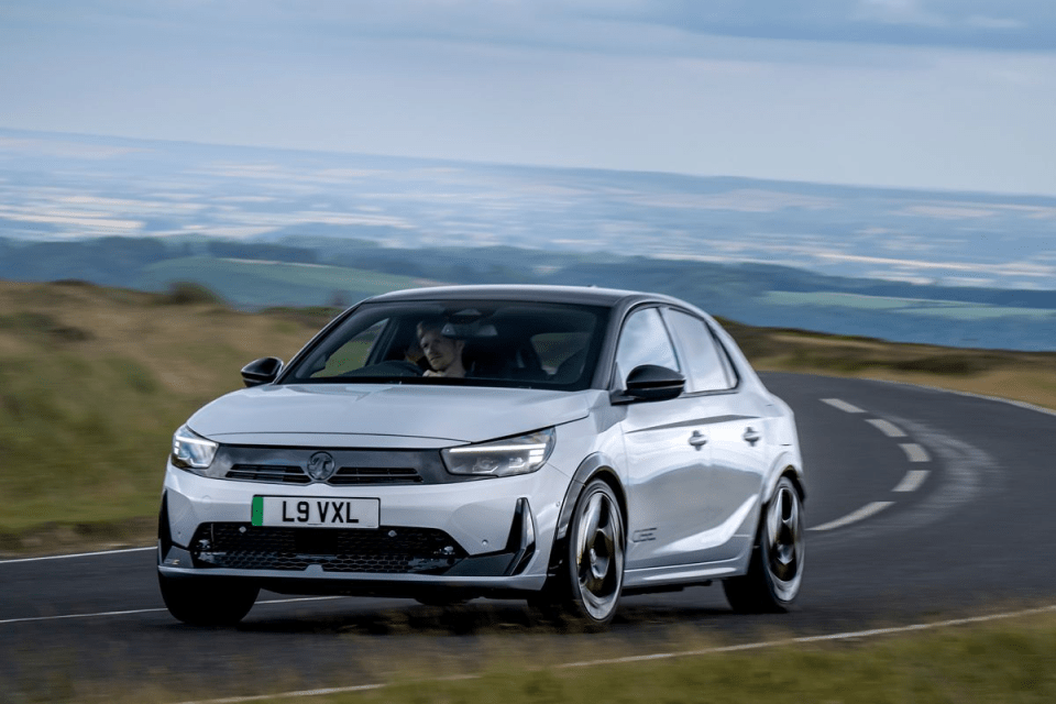 Silver Vauxhall Corsa driving on a winding road with a scenic, hilly landscape in the background.