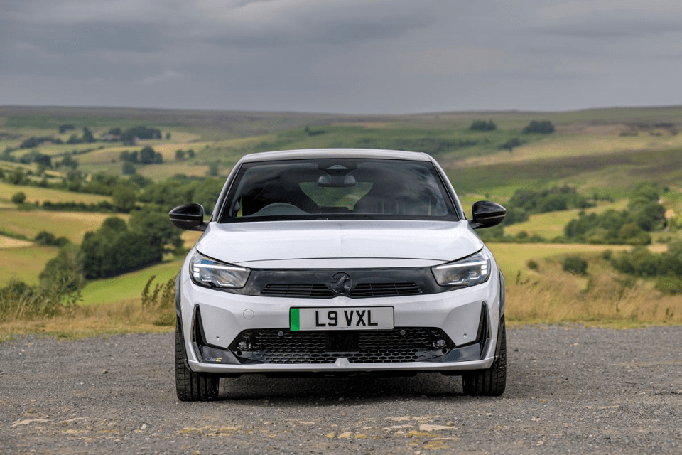 Front view of a white Vauxhall Corsa parked on a gravel road with rolling green hills and a cloudy sky in the background.
