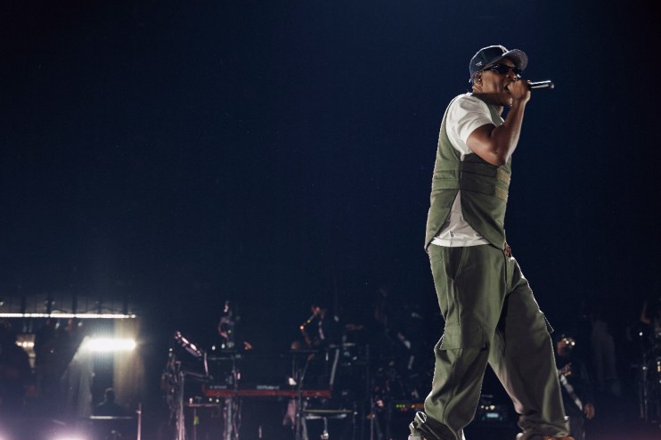 Jay-Z performing on stage at Tottenham Hotspur Stadium, wearing a cap, sunglasses, vest, and cargo pants.