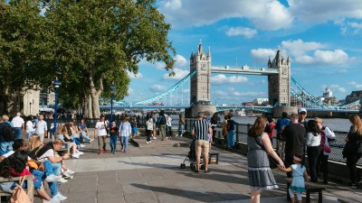 Crowd of tourists and locals walking along the Thames River with the iconic Tower Bridge in London, UK, on a sunny day.