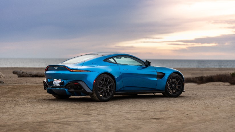 Blue Aston Martin Vantage parked on a sandy beach, ocean and sunset sky in background.