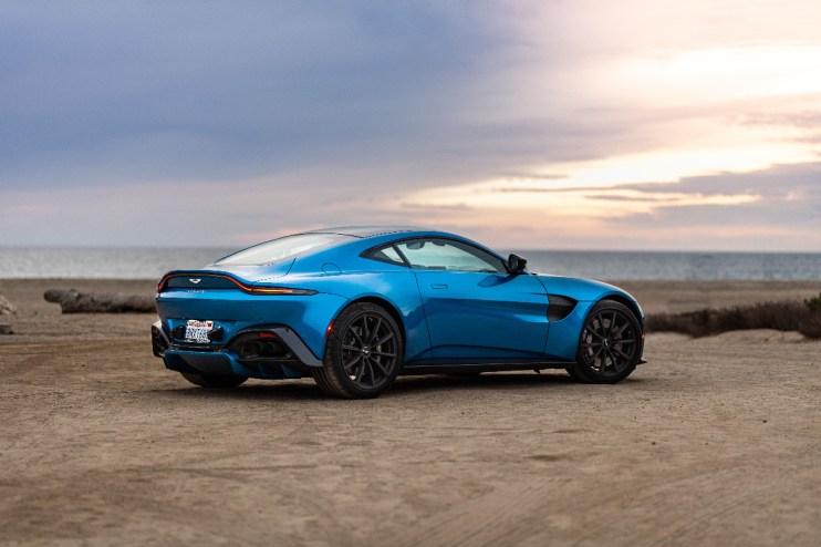 Blue Aston Martin Vantage parked on a sandy beach, ocean and sunset sky in background.