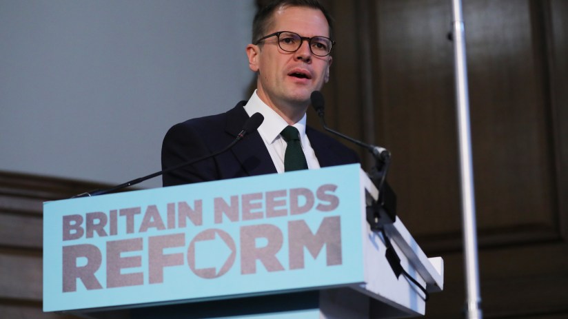 Robert Jenrick speaking at a podium with BRITAIN NEEDS REFORM sign, wearing glasses, suit, and green tie.