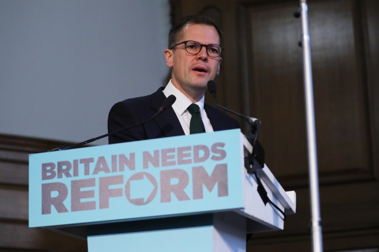 Robert Jenrick speaking at a podium with BRITAIN NEEDS REFORM sign, wearing glasses, suit, and green tie.