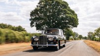 Classic black Rolls-Royce sedan driving on a country road with blurred background foliage and a large tree.